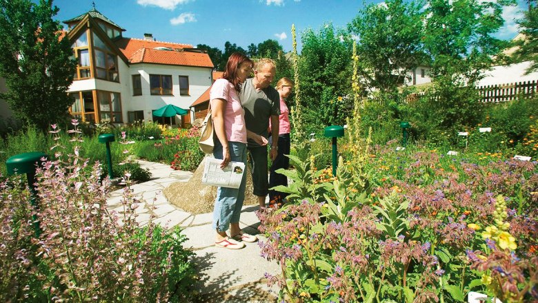 Drei Personen betrachten einen Kr&auml;utergarten vor einem Geb&auml;ude mit roten D&auml;chern.