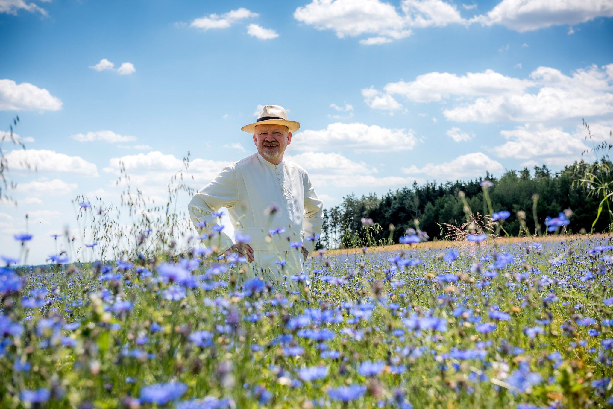 Ein Mann mit Hut steht in einem blühenden Kornblumenfeld unter blauem Himmel.