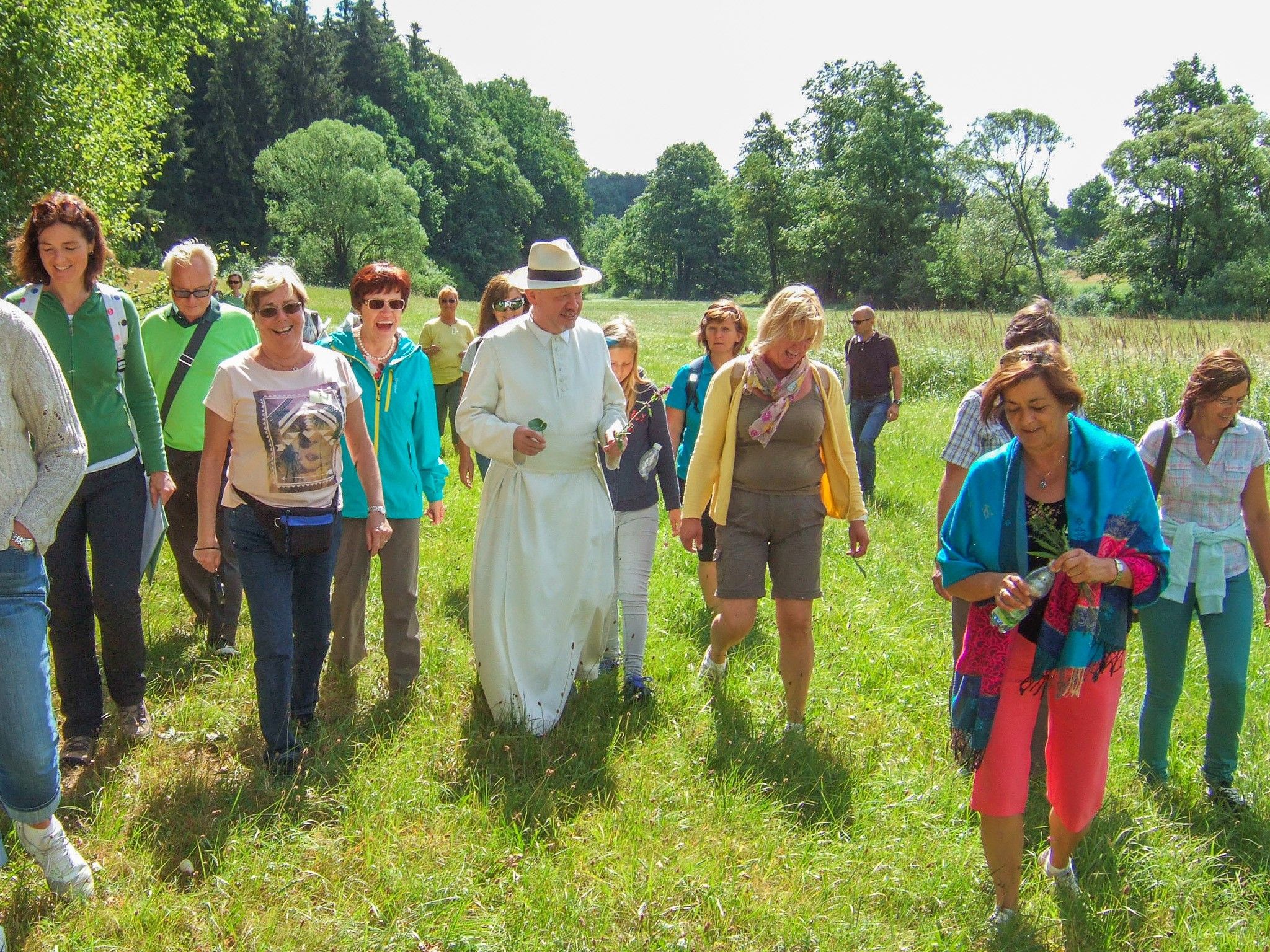 Eine Gruppe von Menschen spaziert über eine Wiese, angeführt von einer Person in weißer Kleidung und Hut. Im Hintergrund sind Bäume zu sehen.